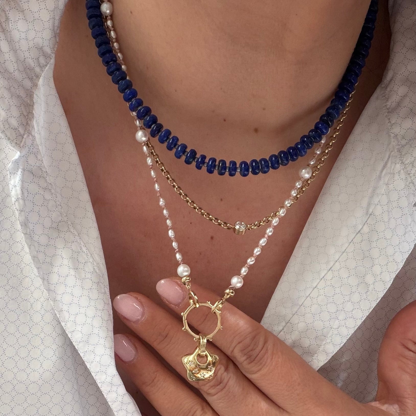 A close-up of a person wearing a layered necklace set. The set includes a short navy blue beaded necklace, a delicate gold chain, and a longer layered necklace with white pearls and a gold ring charm featuring a small gold flower pendant.