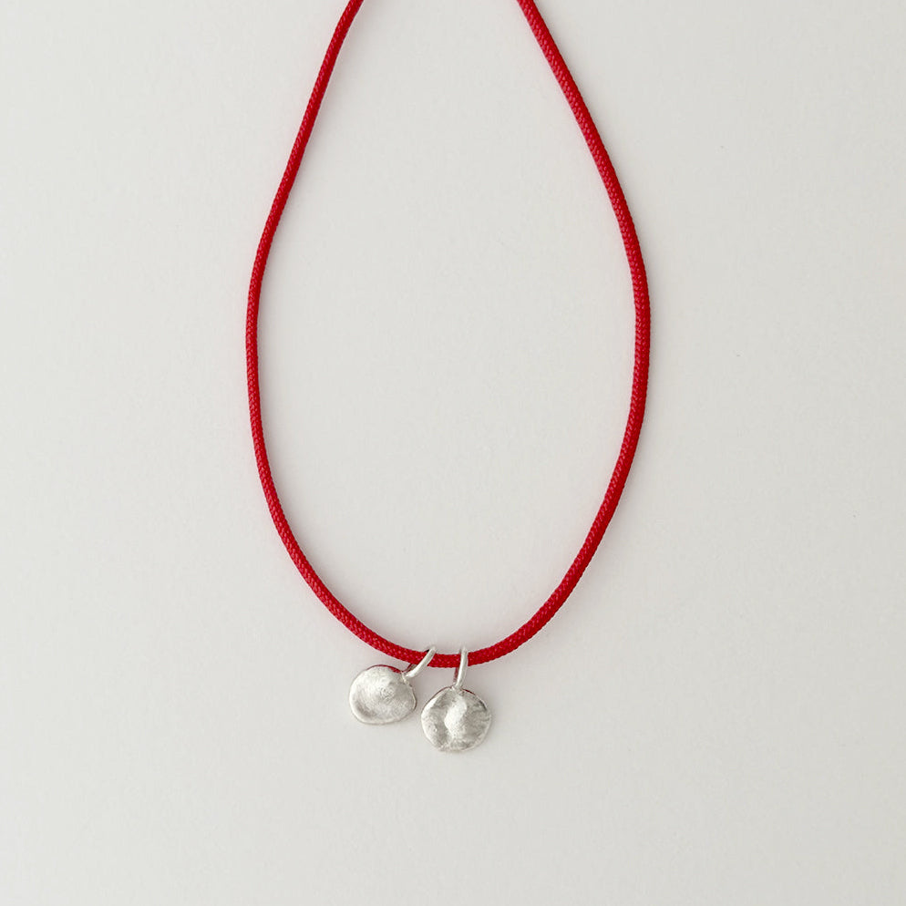 A red cord necklace with two small silver charms, one smooth and one textured, displayed against a plain light gray background.