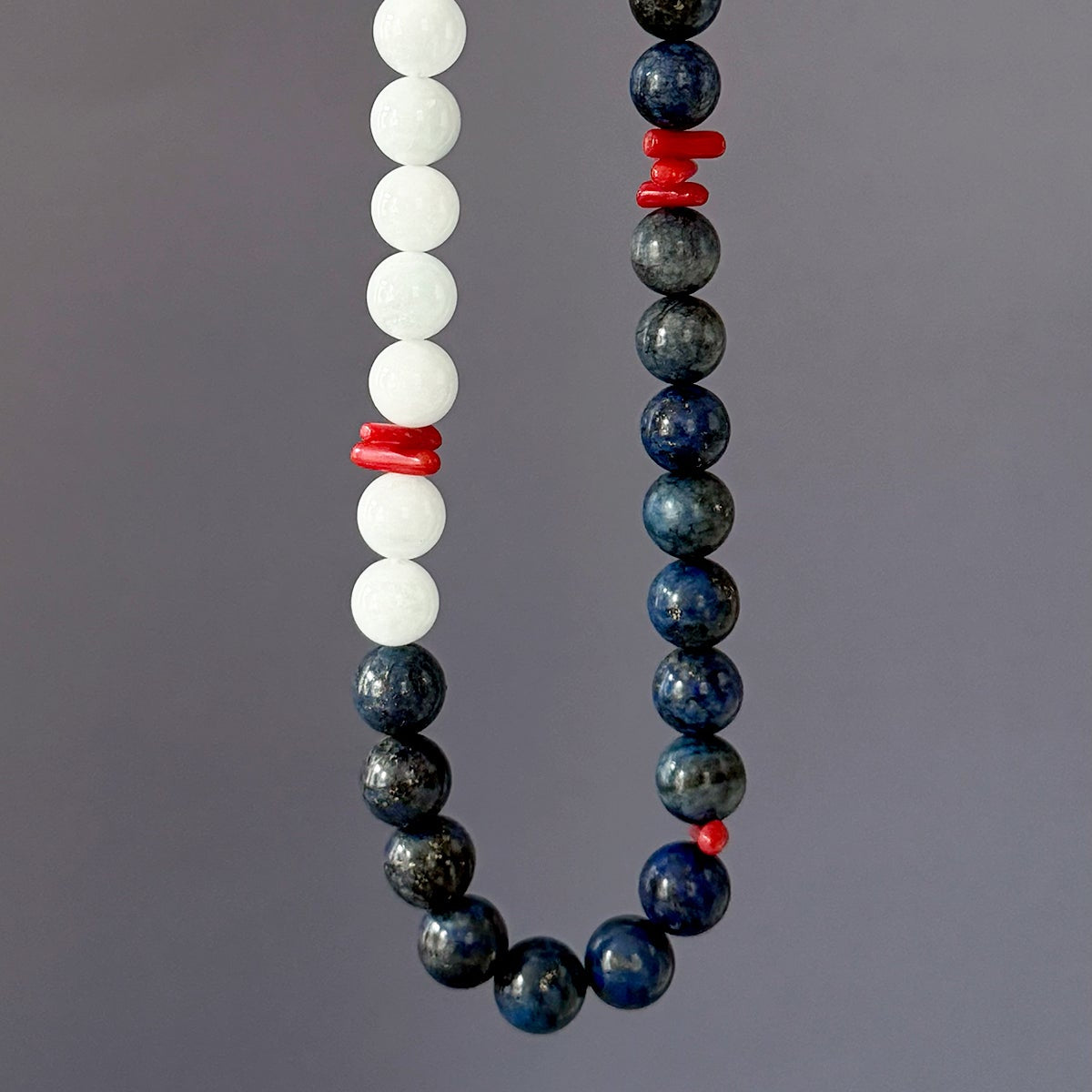 A beaded necklace or strand of round gemstone beads hanging against a plain gray background. One side consists of matte white beads, the other of dark blue-gray beads, and both are separated by small red spacer beads.
