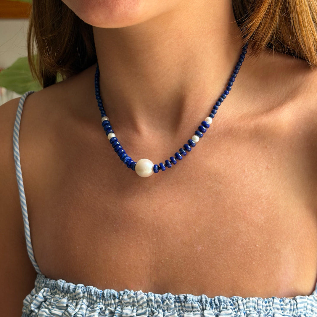 A close-up of a woman wearing a beaded necklace with dark blue beads, small white spacer beads, and a large white pearl at the center, paired with a light blue and white striped top.