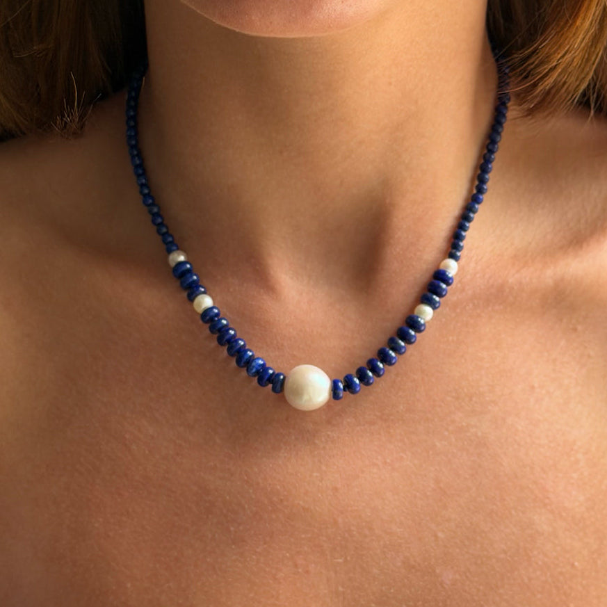 A close-up of a woman wearing a beaded necklace with dark blue beads, small white spacer beads, and a large white oval center bead.
