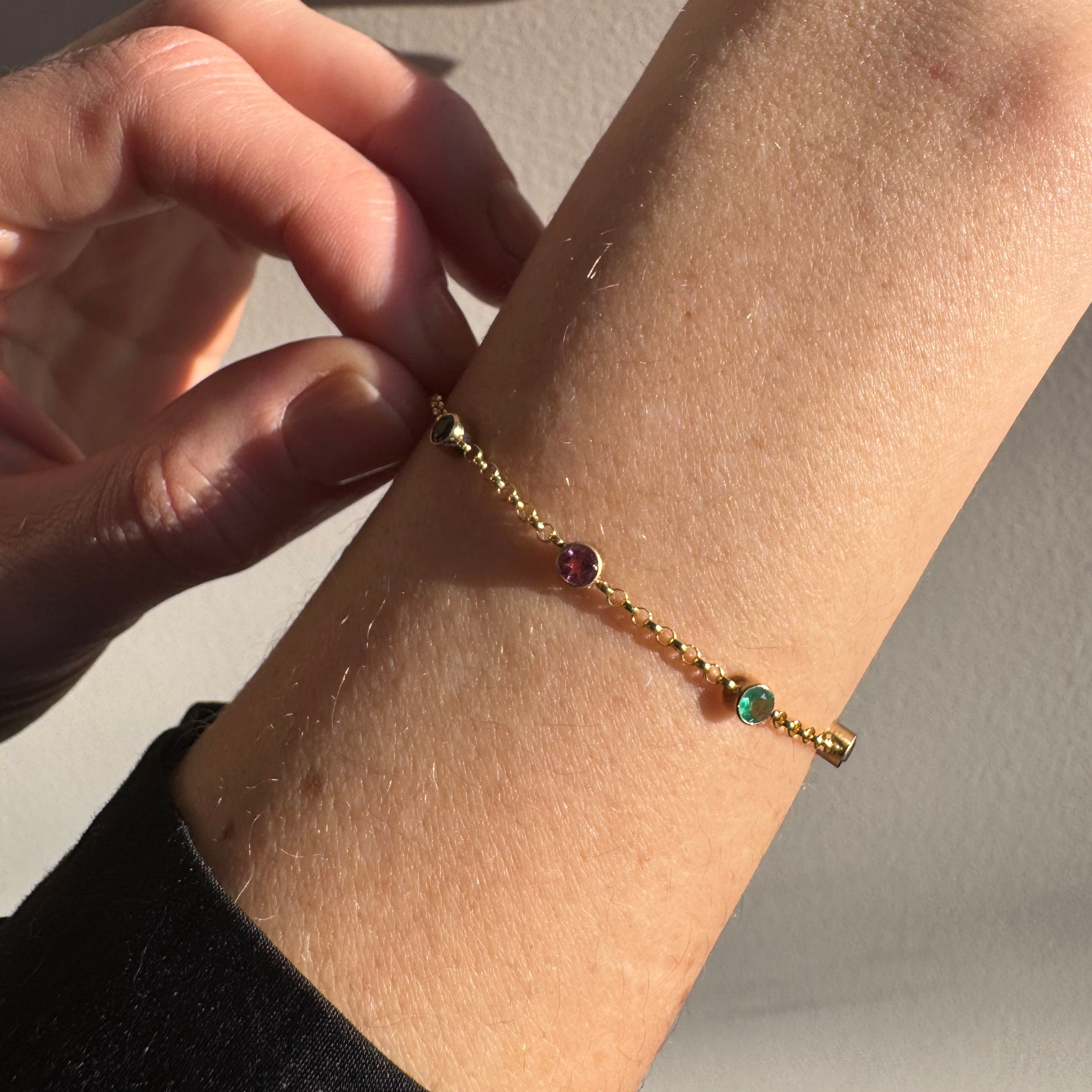A delicate gold chain bracelet with three small round gemstone beads in green, pink, and red, shown worn on a person's wrist against a neutral light gray background.
